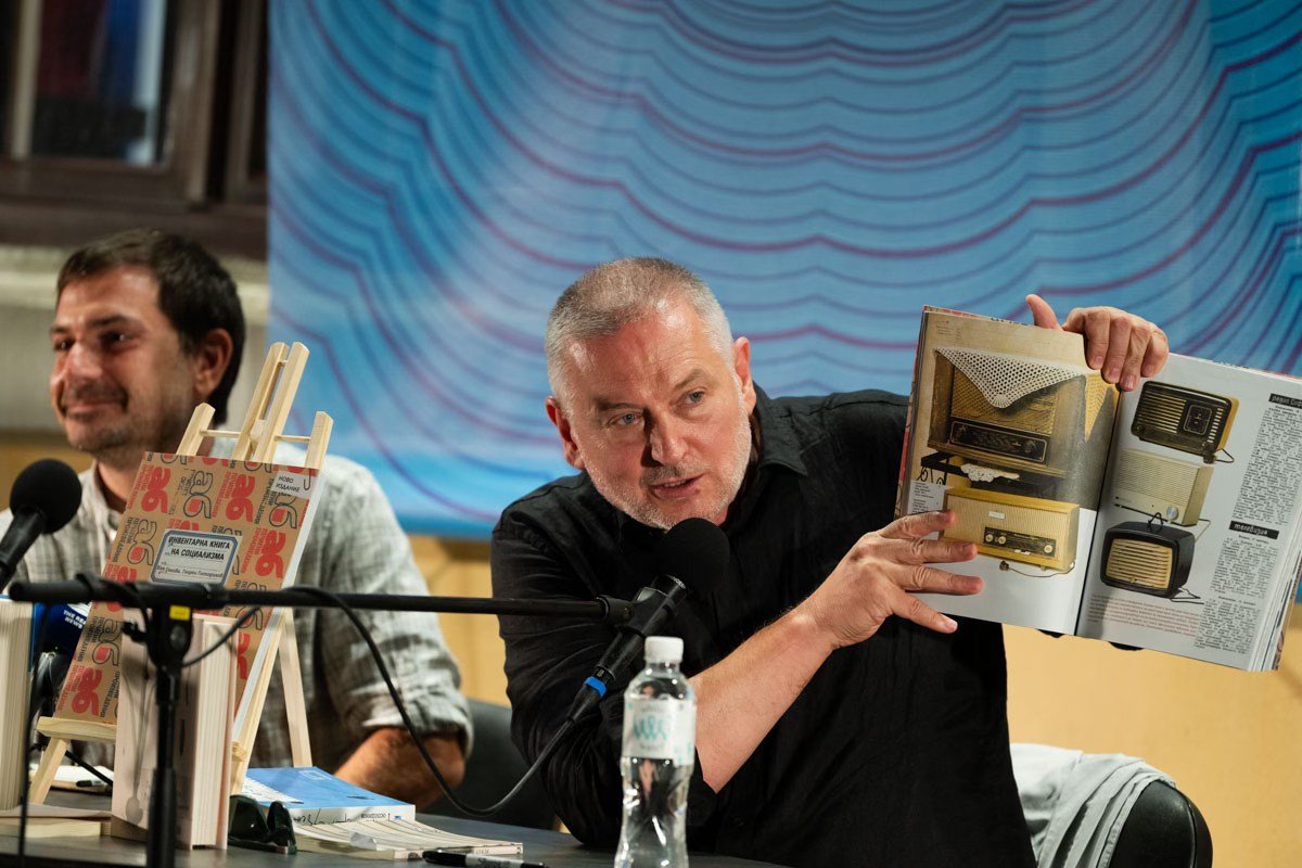 Георги Господинов представя книга по време на литературно събитие, държейки отворено издание пред публика и микрофон.