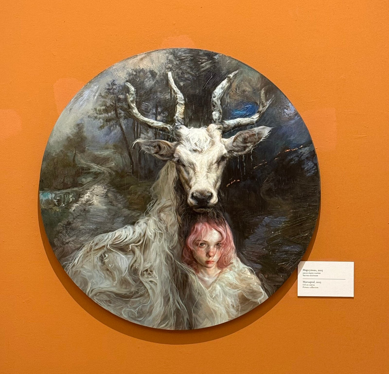 Кръгла картина от изложбата на Гийермо Лорка, изобразяваща момиче с розова коса и бяла коза зад него, поставена на оранжева стена в галер